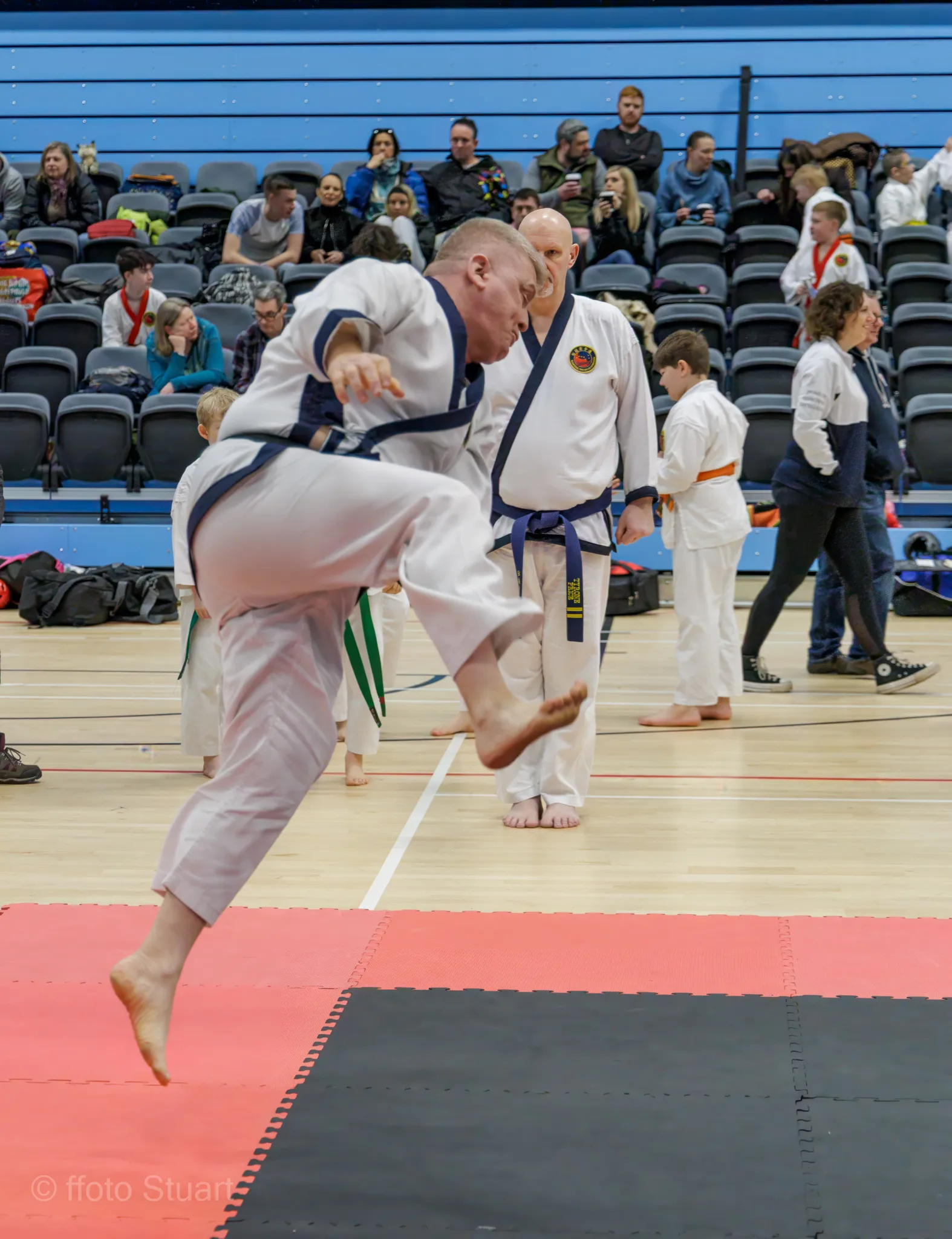 Traditional Tang Soo Do martial artist in uniform performing a jumping kick on a mat, with other participants and seated spectators in a sports hall.