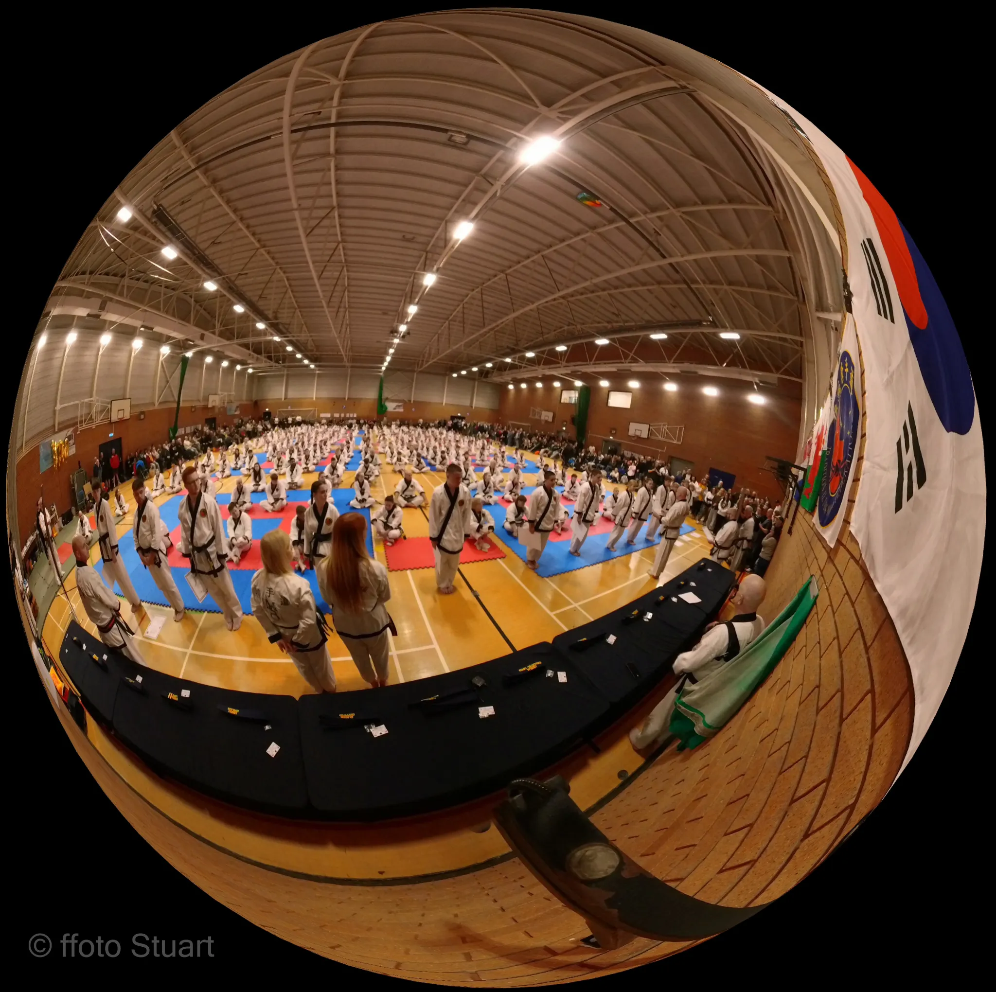 Spherical tiny planet image showing a large Traditional Tang Soo Do grading event in a sports hall, with practitioners in uniform on coloured mats and spectators surrounding the area.