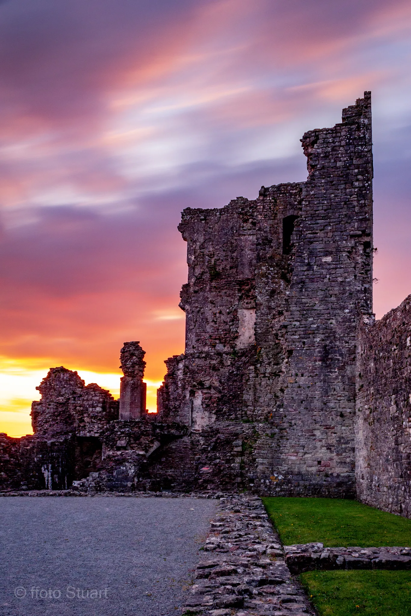 Coity Castle - medieval stone castle with a partially intact tower and surrounding walls, overlooking a quiet courtyard at sunset under streaks of pink, purple and orange clouds.