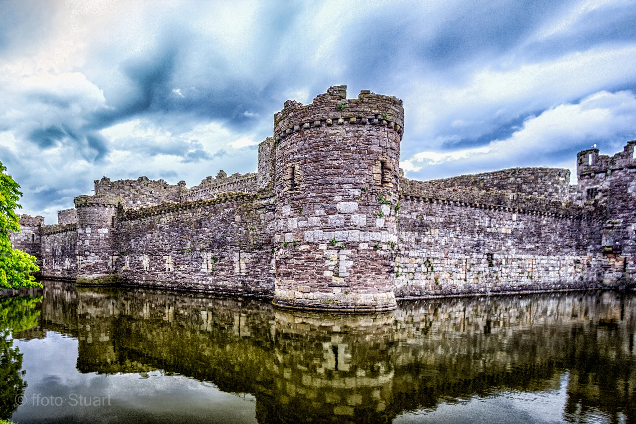 Beaumaris Castle’s concentric stone walls and circular towers mirrored in a still moat, with textured clouds overhead creating a dramatic atmosphere.